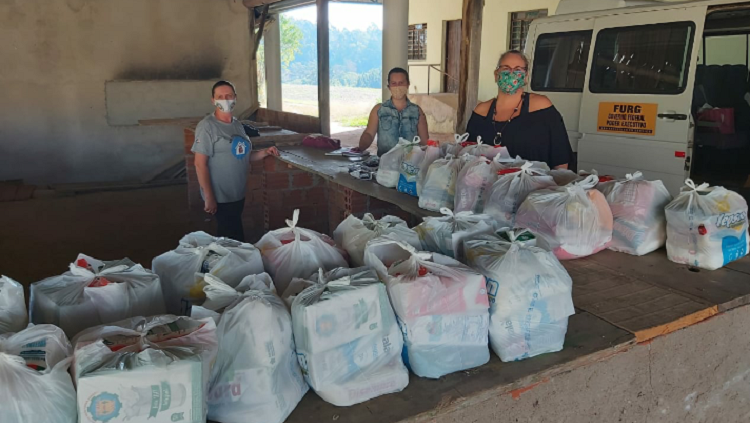 A foto mostra muitas sacolas com alimentos alinhas em cima de um balcão esperando as famílias. Ao fundo, três voluntários da FURG fazendo a distribuição