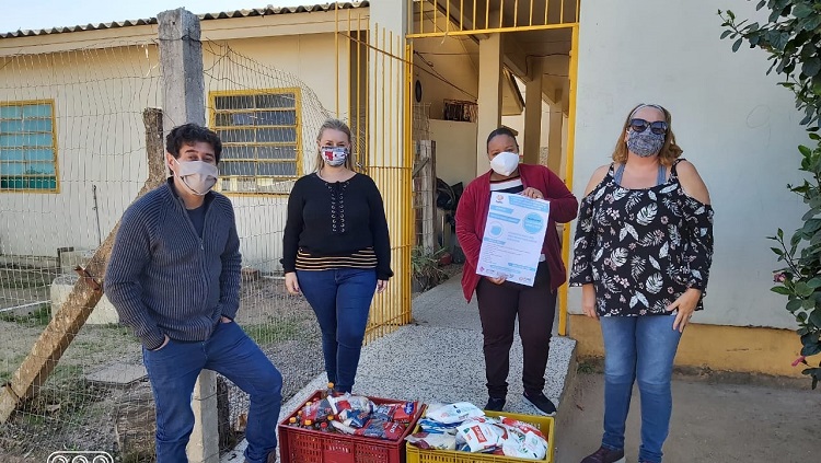 Quatro pessoas aparecem na foto, todas de máscara e uma segurando um cartaz e no chão uma caixa com alimentos