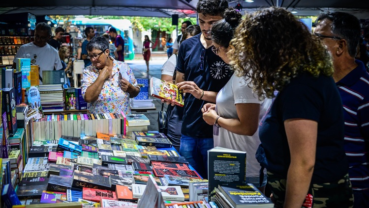 Na foto, um grupo de visitantes conversa entre si em frente a um estande de livros na Feira. Em pé, um homem mostra um livro a duas mulheres.