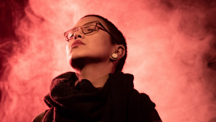 A foto mostra uma mulher em primeiro plano. Ela usa óculos, tem cabelos curtos, raspados, e sua cabeça aponta para cima e à sua direita. Ela está a frente de uma fumaça e iluminação, característicos de palco, em tonalidades de vermelho.