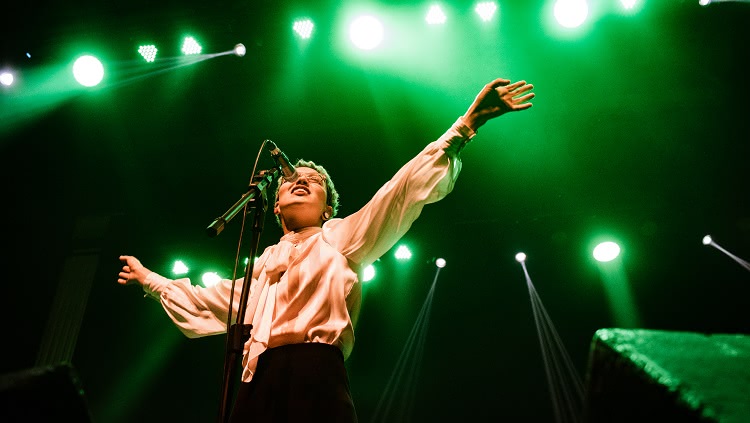 A imagem mostra uma cantora em um palco. A foto foi feita de baixo para cima. Ela usa uma blusa branca e está com os dois braços abertos, cantando diante de um microfone. A foto mostra a iluminação do palco, em tons de verde