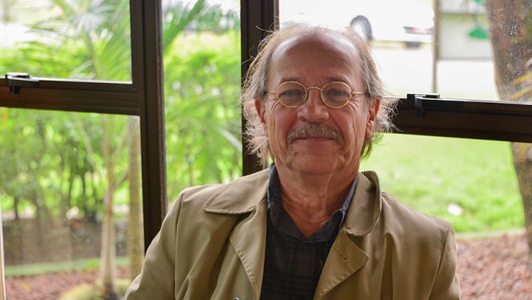 A foto mostra um homem em primeiro plano. Ele tem os cabelos grisalhos nas laterais da cabeça e um pouco de calvície no topo. Ele usa óculos redondos, tem um bigode grisalho e está sorrindo com os lábios fechados. Ele usa um casaco de cor bege e está sentado à frente de uma janela. Ao fundo, podem ser vistas plantas, árvores e um carro desfocado. 