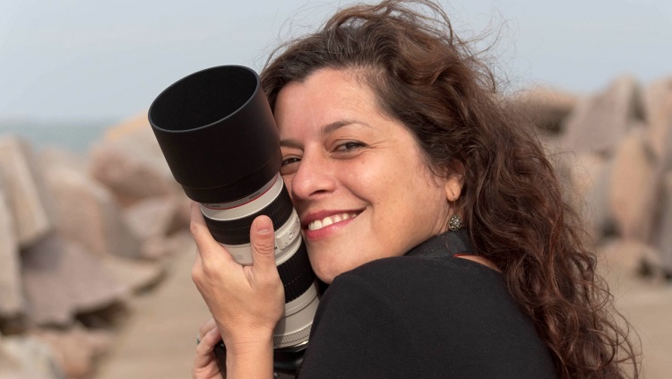 Daniela Delias aparece em primeiro plano. Ela tem cabelos compridos, ondulados, castanhos e está abraçada na lente de uma câmera fotográfica. Ela sorri. Ao fundo é possível ver as pedras dos molhes da barrra desfocadas. 