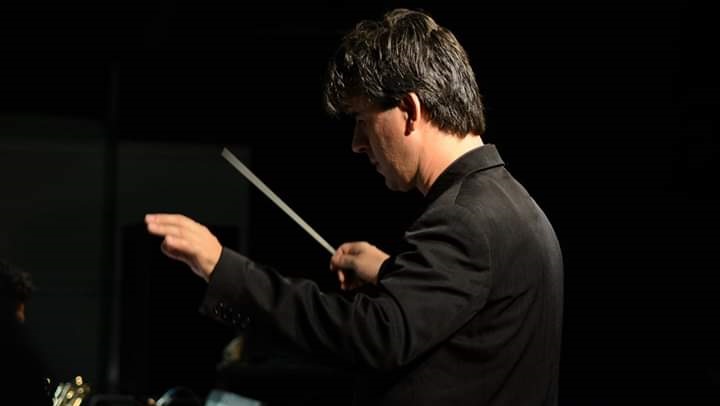A foto mostra um homem de perfil, atuando como maestro. Ele usa um blazer preto. É possível ver seu rosto, concentrado e os braços. O fundo da foto também é preto. 