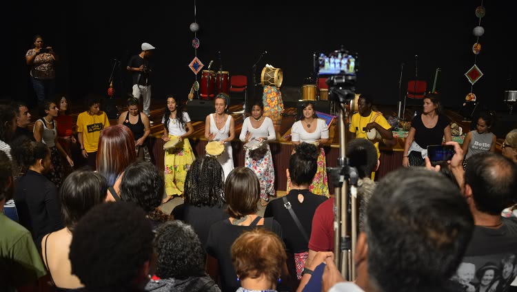 A imagem mostra muitas pessoas. Em frente a um palco, quatro mulheres, que usam saias longas e coloridas, tocam instrumentos de percussão como pandeiros. Há músicos em cima do palco e muitas pessoas aparecem de costas para a foto, porém assistindo às mulheres que tocam. 