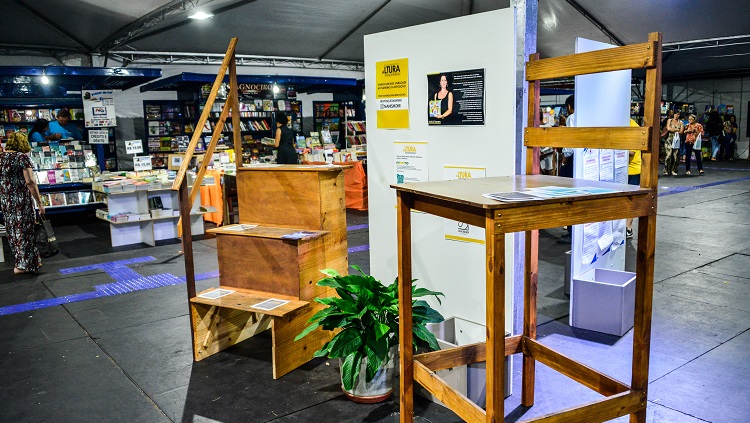 No centro da foto estão duas estruturas de madeira. Uma delas é uma escada de três degraus e corrimão, e a outra é uma cadeira alta. Entre elas, há um painel de cor branca com cartazes e um vaso com plantas. Ao fundo do cenário, pode-se ver alguns estandes da Feira do Livro da FURG.