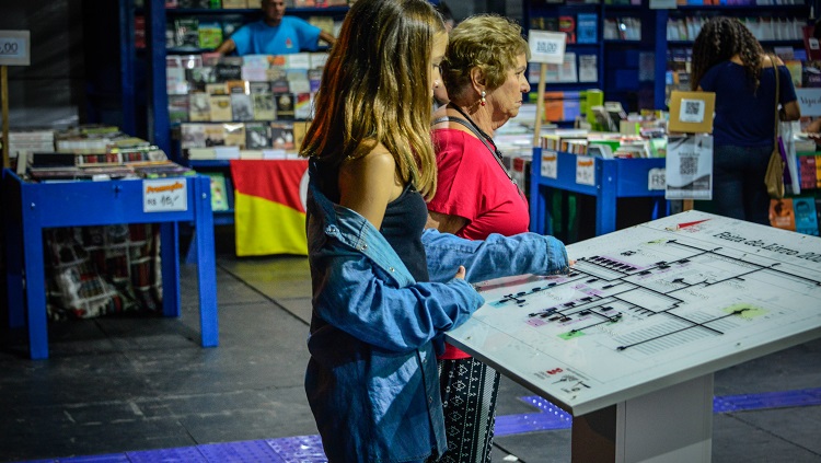 Uma jovem de cabelos claros e jaqueta jeans toca com a mão esquerda o mapa tátil da Feira do Livro da FURG. Ao fundo da foto, em desfoque, estão alguns estandes de livros.