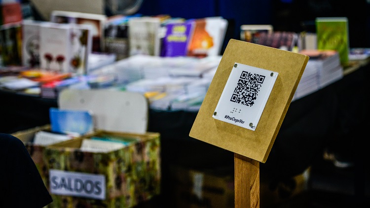 Um totem de madeira exibe uma placa que exibe um QR Code e, abaixo, a hashtag #PraCegoVer. Ao fundo, em desfoque, um estande de livros e uma caixa onde se lê a palavra 