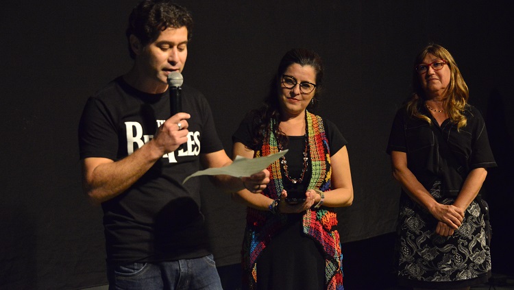 Na foto, o pró-reitor Daniel Prado segura um microfone e lê um papel. Ao lado dele, a patrona e a reitora da FURG.