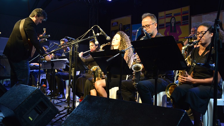 Na foto, parte da Big Band da FURG sentados tocando instrumentos de sopro e o regente da banda.