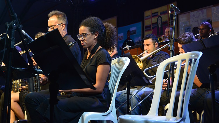 Na foto, parte da Big Band da FURG sentados tocando instrumentos de sopro e o regente da banda.