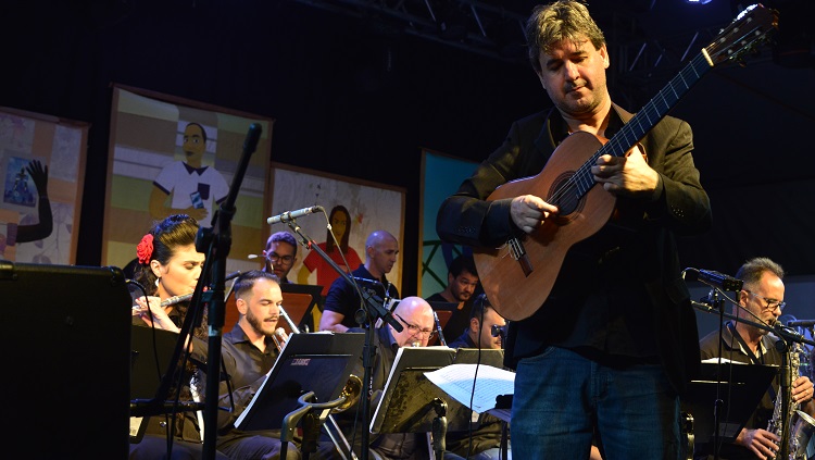 A foto mostra um músico tocando violão e a Big Band ao fundo.