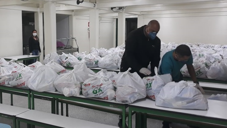 A foto mostra o espaço de um refeitório. Em duas mesas compridas, estão dispostas muitas sacolas de supermercados, cheias. Dois homens, usando máscaras faciais e luvas, aparecem na foto, organizando as sacolas. Eles estão bem à direita da imagem. Na esquerda da foto, ao fundo do refeitório, aparece uma mulher em pé, também com luva e máscara. 