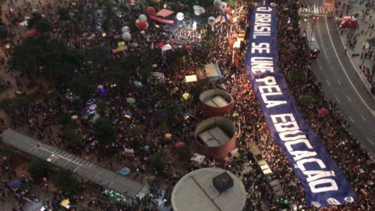 A foto mostra uma multidão em protesto. É uma foto feita à noite, tirada de um ponto alto, que mostra a multidão em um parque ou praça de um ângulo superior. À direita da imagem lê-se numa grande faixa: O Brasil se une pela educação.