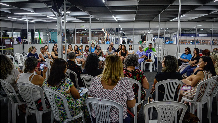 Na imagem aparece um grande grupo de pessoas sentadas em círculo. Elas estão em cadeiras brancas em um ambiente que é cercado por paredes provisórias, com a parte inferior em material branco e a parte superior em vidro. 