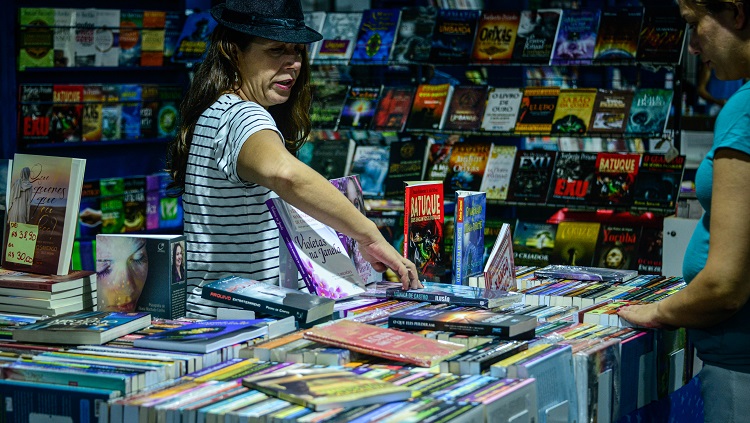 Na imagem aparece uma banca de livros. A foto mostra uma mulher dentro da banca. Ela usa um chapéu e aponta para um livro enquanto outra mulher, do lado de fora da banca, a observa, com as mãos sobre os livros. 