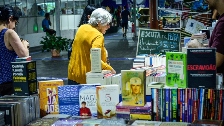 Em primeiro plano aparecem capas de livros desfocadas. Ao fundo, uma senhora de cabelos brancos, que usa uma blusa amarela, olha os livros. Há um pequeno quadro com o escrito em giz: escuta na feira, atrás dos livros. 