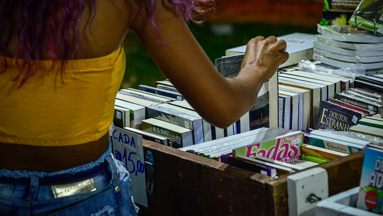 Uma jovem mulher aparece de costas na foto. A foto a enquadra da cintura para cima. Ela usa uma blusa curta e está com cabelos, de cor roxa soltos. Sua posição na foto é à esquerda. À direita aparecem livros de uma banca. A jovem está pegando um livro com a mão. 