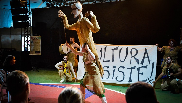 Artistas aparecem em uma performance. Uma mulher manipula um boneco, que está acima de seu corpo. Todos vestem roupas de estopa. Atrás da mulher, três homens e uma mulher, com figurino semelhante, seguram uma faixa em que consta o texto: cultura resiste. 