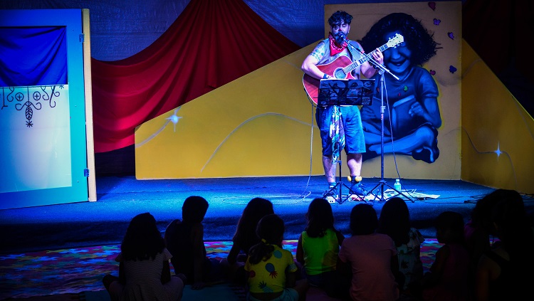 Em um palco com um fundo colorido, com cortinas vermelhas e amarelas, está um palhaço. Ele toca violão e olha para o público, que aparece de costas na foto, assistindo ao show. 