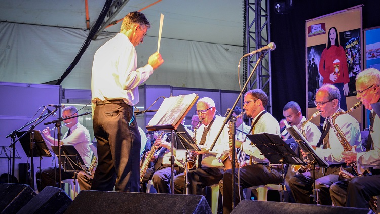 A foto mostra muitos músicos homens em um palco. Eles tocam instrumentos de sopro. À frente deles, de costas para a foto, está um maestro, que gesticula com a mão ao alto, segurando uma batuta. 