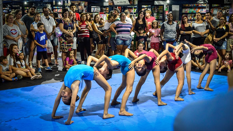 Seis meninas aparecem na foto fazendo uma acrobacia ginástica. Cada uma vira o corpo em direção à que está atrás de si, segurando a cintura da colega. Elas usam malhas de ginástica e estão sobre um fundo azul. Há bastante pessoas assistindo à apresentação. 