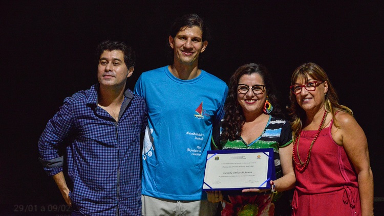 Quatro pessoas posam para a foto. Elas estão à frente de um fundo preto. À esquerda, o pró-reitor Daniel Prado. Ao lado dele, o vice-reitor Danilo Giroldo. Ao lado, a patrona da Feira, Daniela Delias. Ela segura uma placa de homenagem, que mostra à foto. E finalmente, à direita, a reitora Cleuza Dias. 