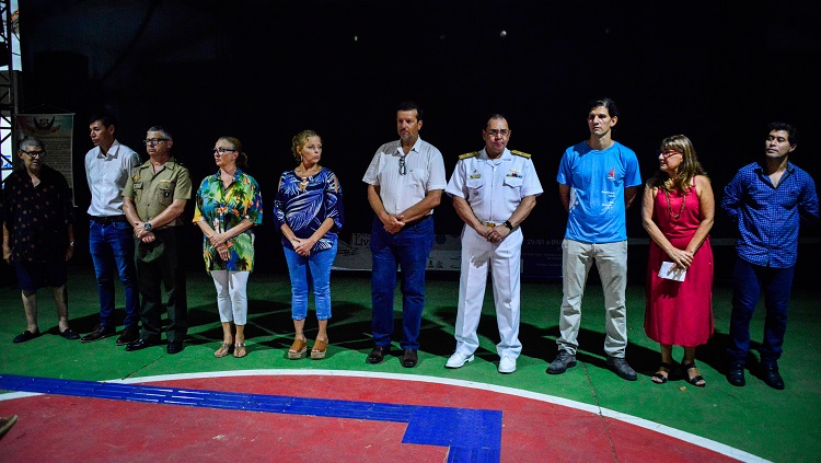 A imagem mostra um grupo de pessoas dispostas lado a lado. São dez pessoas, entre homens e mulheres. Eles estão à frente de um palco que está com as cortinas cerradas. 