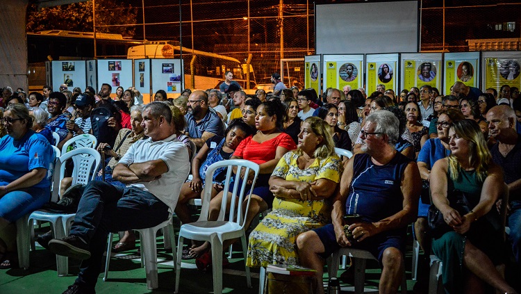 Na foto aparecem pessoas sentadas, é o público da abertura da feira. As pessoas estão sentadas em cadeiras brancas e olham para a frente. O espaço é de uma quadra esportiva e a cena se passa à noite. Ao fundo há pôsteres com imagens e textos, que não são legíveis na foto. 