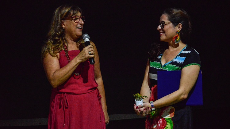 Duas mulheres aparecem na foto sobre um fundo escuro. Elas estão se olhando. À esquerda, a reitora Cleuza. Ela fala, com o microfone à mão e sorri. À direita, a patrona da feira Daniela Delias. Ela também sorri enquanto olha para a reitora. Ela tem uma pasta embaixo do braço e segura uma flor com as duas mãos em frente ao corpo. 