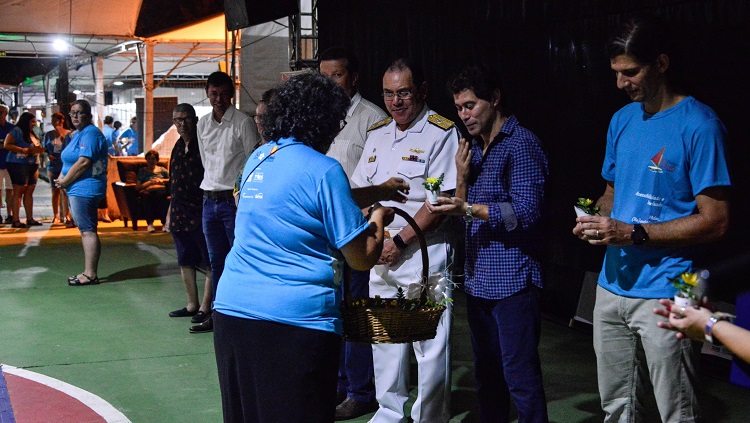Uma mulher aparece de costas. Ela usa a camiseta azul da feira do livro e está entregando flores de uma cesta às autoridades que estão na abertura da feira do livro. 