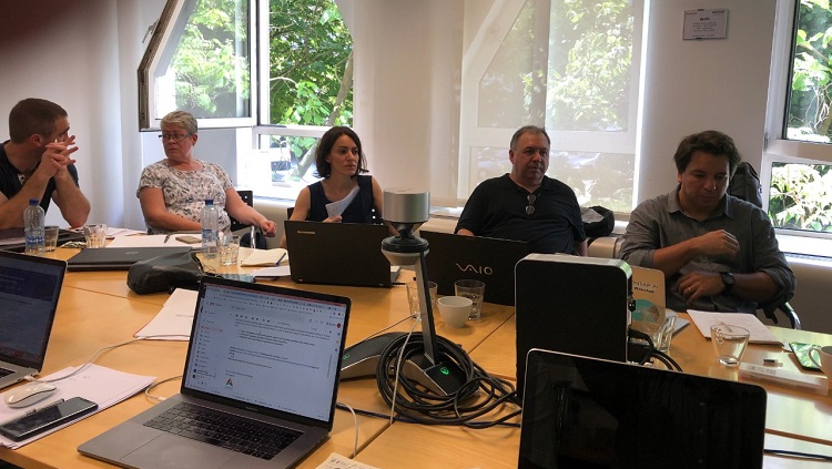 A foto mostra pessoas reunidas em volta de uma mesa, em reunião. Todas estão com computadores notebook abertos à sua frente. Aparecem, à esquerda, um homem, ao lado dele duas mulheres e, ao lado delas, dois homens. Dá para ver na foto que há mais pessoas na reunião além das que aparecem no enquadramento da foto, pois há mais computadores abertos e não aparece a mesa inteira. Atrás das pessoas que aparecem na foto há grandes janelas abertas e dá para ver árvores ao lado de fora