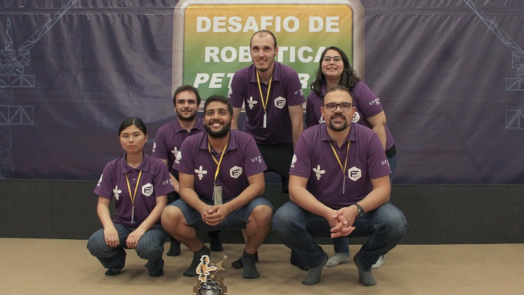 Foto de uma equipe de 6 pessoas. São 4 homens e duas mulheres. Todos vestem camiseta na cor roxa e usam crachá com cordão amarelo. Alguns estão agachados, outros em pé, reunidos para a foto. à frente deles, no chão, há um troféu. No fundo aparece escrito "Desafio de robótica Petrobras"