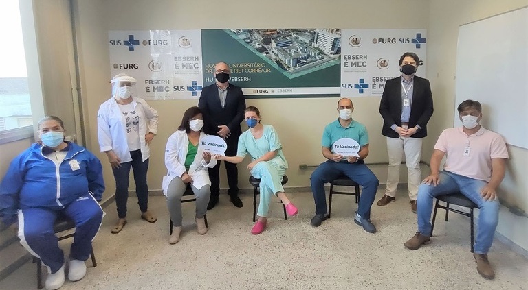 Na foto, aparecem oito pessoas, entre homens e mulheres, 5 estão sentados. Todos usam máscaras de proteção para o coronavírus e estão em um local em que ao fundo há um mural com o nome do HU-FURG Ebserh e uma foto do hospital. Duas pessoas sentadas seguram placas eme formato de balão de texto em que está escrito: "Tô vacinado". 