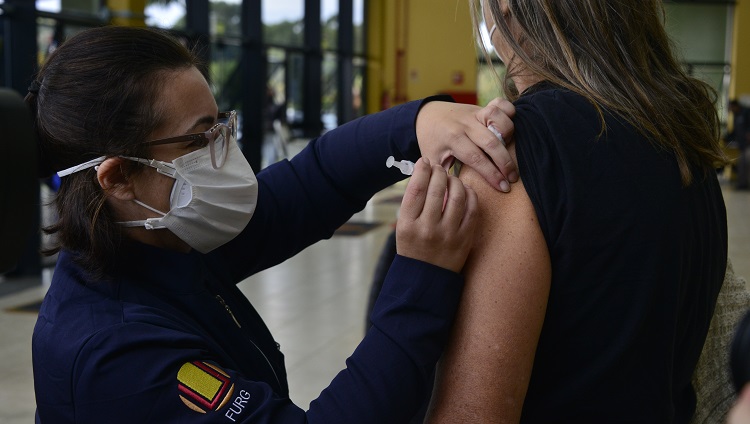 A foto mostra uma profissional de saúde, com máscara, vacinando uma mulher, que aparece de costas na imagem.