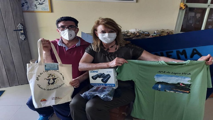 A imagem ostra um homem e uma mulher sentados, usando máscaras de proteção contra a Covid-19. Ambos mostram uma bolsa reciclável, camiseta e binóculos, prêmios que ganharam no concurso.