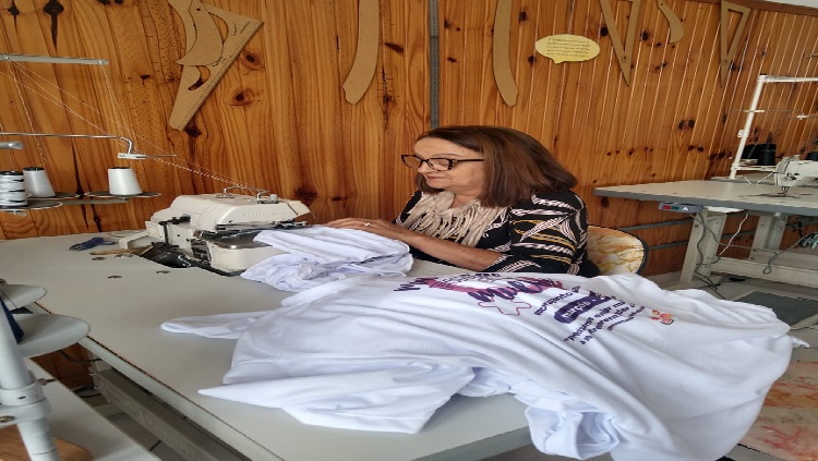 A imagem mostra uma mulher sentada de cabelos compridos, atrás de uma mesa confeccionando camisetas