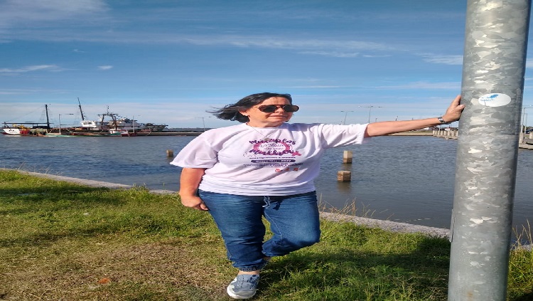 A imagem mostra uma mulher em pé, vestindo camiseta branca e calça jeans, tendo ao fundo a lagoa dos patos.