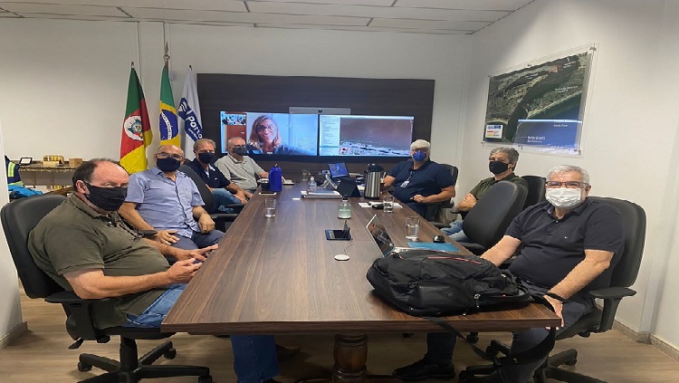 A imagem mostra uma mesa grande com pessoas sentadas a volta dela, dentro de uma sala. Ao fundo da imagem aparece a projeção de um rosto.