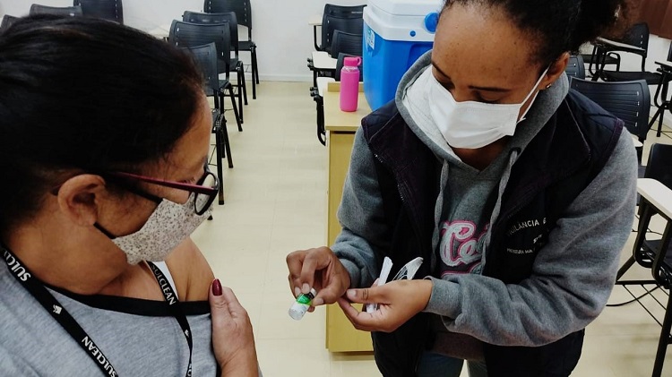 Na fotografia aparecem duas mulheres em uma sala de aula, uma delas com a gola da blusa baixa, expondo seu ombro, e a outra mostra a ela uma ampola de vacina. Elas estão em primeiro plano na foto. A mulher que está com o ombro à mostra para a vacina é parda, tem os cabelos pretos amarrados, usa óculos, uma máscara de pano está com uma camiseta cinza e usa um crachá da empresa sulclean. A mulher que mostra a ampola é negra, tem os cabelos pretos cacheados presos, usa máscara cirúrgica branca e vestes de moletom cinza, com um colete preto sobre a roupa. Atrás delas, é possível ver uma caixa térmica na cor azul. 