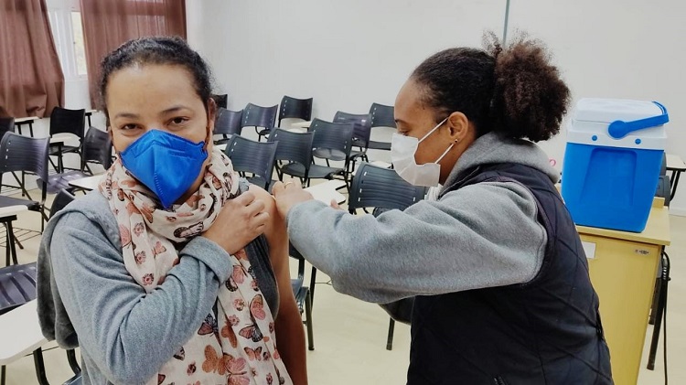 Na fotografia aparecem duas mulheres, uma vacinando e a outra sendo vacinada. Elas estão num espaço de uma sala de aula. A mulher que recebe a vacina está olhando de frente para a foto. Ela usa máscara padrão pff-2 na cor azul, está com os cabelos escuros amarrados e usa um lenço no pescoço. A mulher que aplica a vacina é negra, tem os cabelos crespos presos e usa uma máscara cirúrgica branca. Ela veste um jaleco preto sobre um moletom cinza. Ambas aparecem na foto da cintura para cima. Ao fundo é possível ver cadeiras na sala de aula e uma caixa térmica azul sobre uma mesa amarela. 