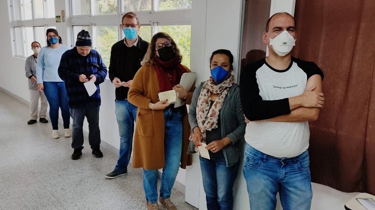 A foto mostra uma fila de pessoas em um espaço que parece ser um corredor, com muitas janelas. Em primeiro plano, na direita da foto, aparece um homem branco de cabelos raspados com máscara branca. Ele usa uma camiseta de manga comprida e, em um dos braços, a manga está levantada, com o braço à mostra. Atrás dele, uma mulher parda, com cabelos pretos presos, máscara azul, roupas de inverno e um lenço estampado no pescoço. Depois, atrás dela, uma mulher branca, de cabelos claros e soltos, segura alguns papéis. Ela usa um casaco comprido, na cor marrom e está com máscara preta. Atrás dela, há dois homens; um loiro e alto com máscara azul e outra mais baixo, que usa touca de inverno e está sem máscara. Atrás deles, duas mulheres encerram a fila, bem na direita da foto. Uma alta, com cabelos castanhos soltos e máscara azul, outra mais baixa, com uniforme da empresa Sulclean, cabelos escuros presos e máscara branca. 